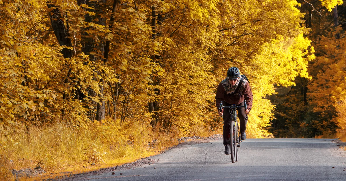 Ciclista con casco rodando por una carretera secundaria entre árboles de otoño