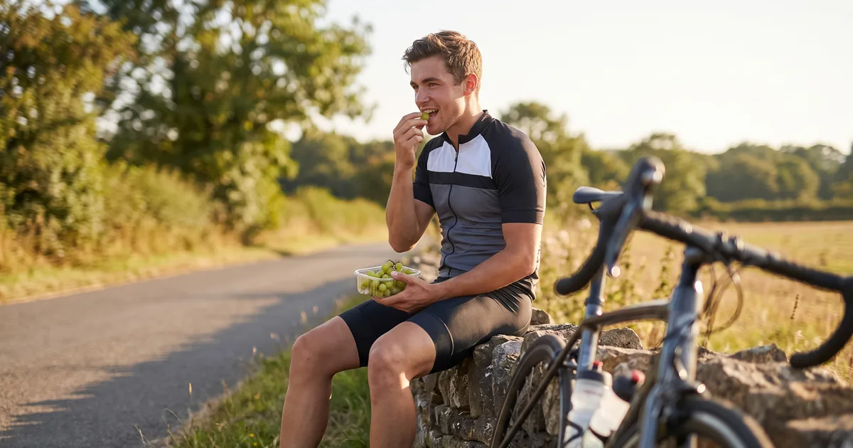 Ciclista sentado en un muro de piedra junto a la carretera comiendo uvas, con su bici de carretera apoyada al lado