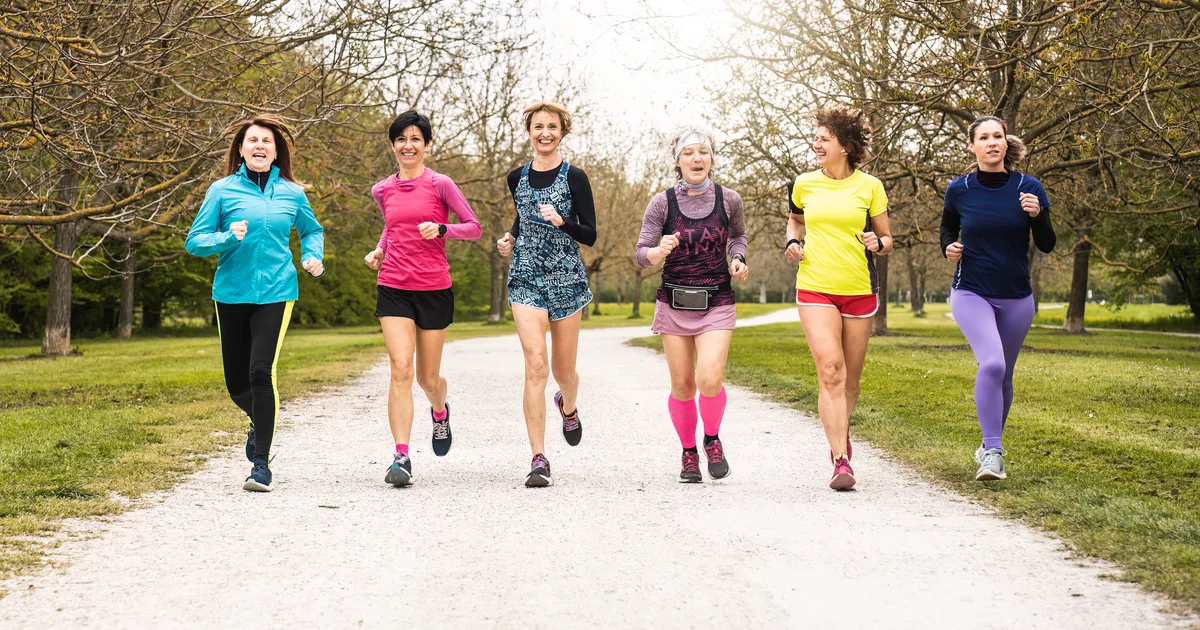 Grupo de seis mujeres de distintas edades corriendo juntas y sonriendo por un camino de un parque
